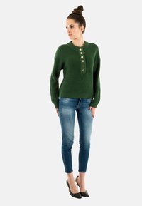 Pull vert côtelé avec col rond et cinq boutons dorés, associé à un jean skinny bleu et des talons noirs pointus.