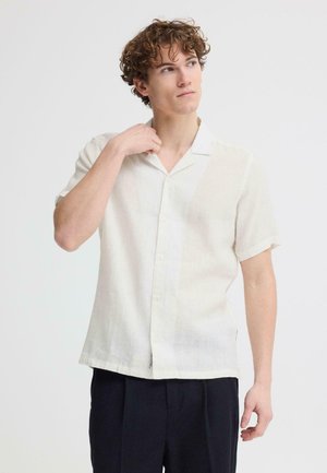 Młody mężczyzna o kręconych włosach, ubrany w białą koszulę na krótki rękaw i ciemne spodnie, poprawiający kołnierzyk z neutralnym wyrazem twarzy.