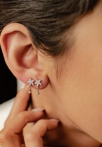 Orecchini a forma di fiore, realizzati in argento, con charm pendenti, caratterizzati da cristalli trasparenti e dettagli intricati nei petali, indossati su un orecchio umano.