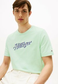 Camiseta verde menta de algodón con un logotipo bordado azul "J. Hilfiger". Mangas cortas y corte estándar con un detalle del logotipo en el costado.
