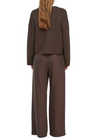 Woman with long brown hair wearing loose, dark brown long-sleeve top and wide-leg pants, standing facing away on white background.