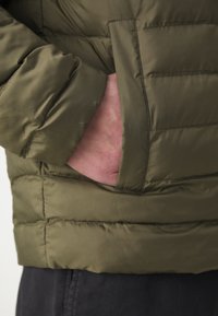 Main insérée dans la poche latérale d'une veste matelassée vert olive, avec un pantalon noir visible en dessous.