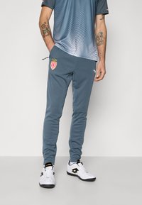 Pantalons de sport gris à coupe slim, ornés d'un logo brodé de Monaco, avec des poches latérales et une taille à cordon, associés à des baskets blanches.