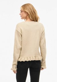 Beige strikkegenser med rysjekant nederst og på mansjettene. Har en ledig passform med rund hals og lange ermer.