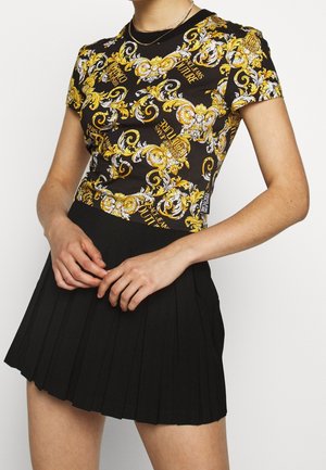 Personne portant un T-shirt moulant noir et or à motifs ornés avec le texte "Versace Jeans Couture" et une mini-jupe noire plissée.