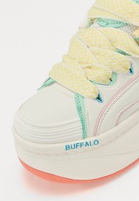 Bijela platforma tenisica s višebojnim naglascima, sa žutim čipkastim vezicama u obliku cik-caka, teksturiranim premazom i plavim "BUFFALO" logotipom sa strane.