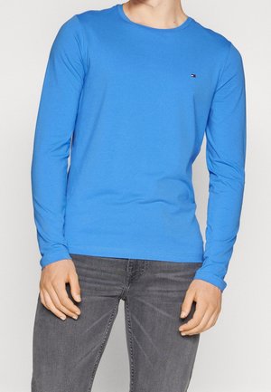 Homme portant une chemise à manches longues bleu vif et un jean gris foncé, debout, mains détendues le long du corps.
