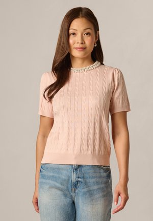 Woman with long brown hair wearing a pink short-sleeve cable knit top with pearl necklace detail and light blue denim jeans.