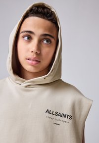 Top beige senza maniche con cappuccio, logo frontale con la scritta "ALLSAINTS" e testo in giapponese. Tessuto liscio e design casual.