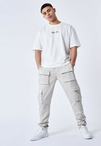 T-shirt bianca a maniche corte con logo nero; pantaloni cargo beige chiaro con molte tasche frontali; sneakers bianche con lacci.