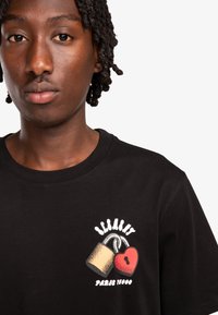Zwarte katoenen T-shirt met grafisch ontwerp van een gouden slot en een rood hart, met daaronder de tekst "Paris 1980" in het wit. Ronde hals.