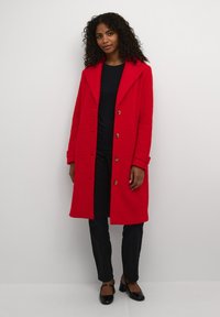 Rode textuur overjas met een reverskraag, zes knoopsluitingen en opgerolde mouwen, gedragen over een zwarte top en zwarte broek.