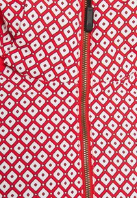Tejido de patrón rojo y blanco con formas de diamante, con un cierre dorado visible y textura suave. El diseño es geométrico y colorido.