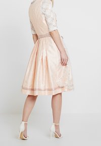 Robe rose blush avec manches en dentelle, corsage ajusté et jupe évasée. Mise en valeur par une ceinture ton sur ton et une texture de tissu complexe.