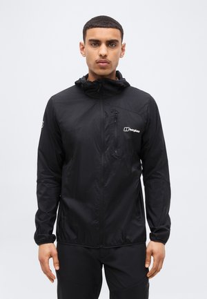 Νέος άνδρας που φοράει μαύρο φούτερ Berghaus με κουκούλα και φερμουάρ στο στήθος, στέκεται μπροστά σε απλό φωτεινό φόντο.