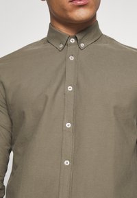 Homme portant une chemise olive boutonnée avec col boutonné, cadré au menton et aux épaules, fond neutre.