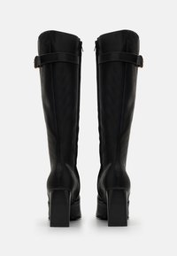 Botas de cuero negro hasta la rodilla con un tacón cuadrado, cierre de cremallera lateral y una correa decorativa alrededor del tubo superior. Textura suave.