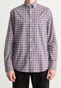 Mann in einem langärmligen, knopfbaren Hemd mit rot, grau und weißem Karo-Muster und schwarzen Hosen, der vor einem neutralen Hintergrund steht.