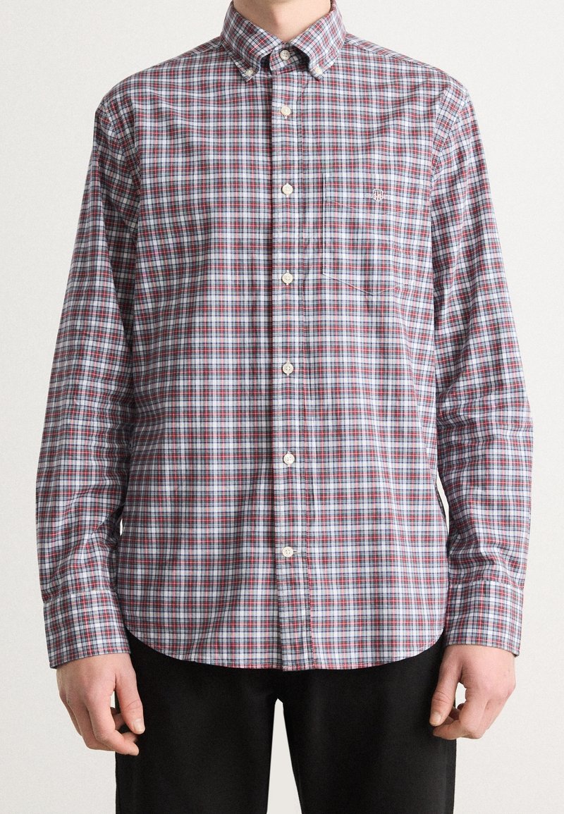 Mann in einem langärmligen, knopfbaren Hemd mit rot, grau und weißem Karo-Muster und schwarzen Hosen, der vor einem neutralen Hintergrund steht.