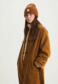 Manteau en laine mélangée marron à coupe décontractée, doté de grands boutons et de poches latérales. Associé à un bonnet côtelé orange et un pull léger.