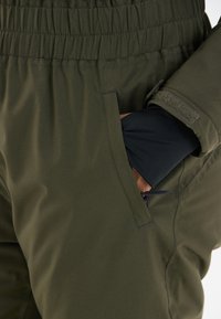 Un pantalon vert olive avec une taille élastique, doté d'une poche latérale. Le matériau est lisse avec un léger éclat et une texture douce.