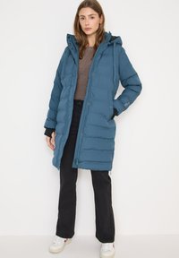 Mujer vestida con un abrigo acolchado azul con capucha sobre un suéter marrón, pantalones negros y zapatillas blancas, de pie contra un fondo blanco liso.