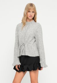 Γκρίζα πλεκτή ζακέτα με στρογγυλή λαιμόκοψη, μακριά μανίκια, ζωνάκι στη μέση και τσακισμένα άκρα στα μανίκια, συνδυασμένη με μια μαύρη φούστα με τσακίσεις.