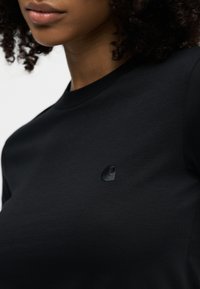 Κλείσιμο σε πρόσωπο που φοράει μαύρο T-shirt με μικρό, μαύρο κεντητό λογότυπο στο στήθος, με φαίνoμενα σγουρά μαλλιά.