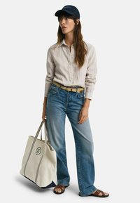 Camisa de lino beige claro de botones, jeans de pierna ancha de mezclilla, cinturón trenzado, gorra marina, sandalias marrones y un gran bolso de lona crema con acentos en azul marino.