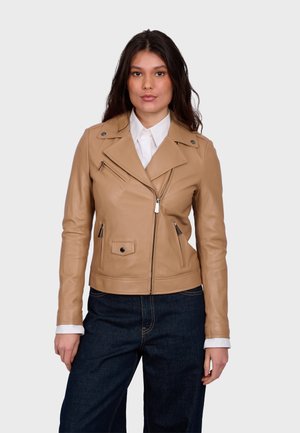 Žena v béžové kožené motorkářské bundě přes bílou košili s límečkem a tmavě modré džíny, stojící na jednoduchém světlejším pozadí.