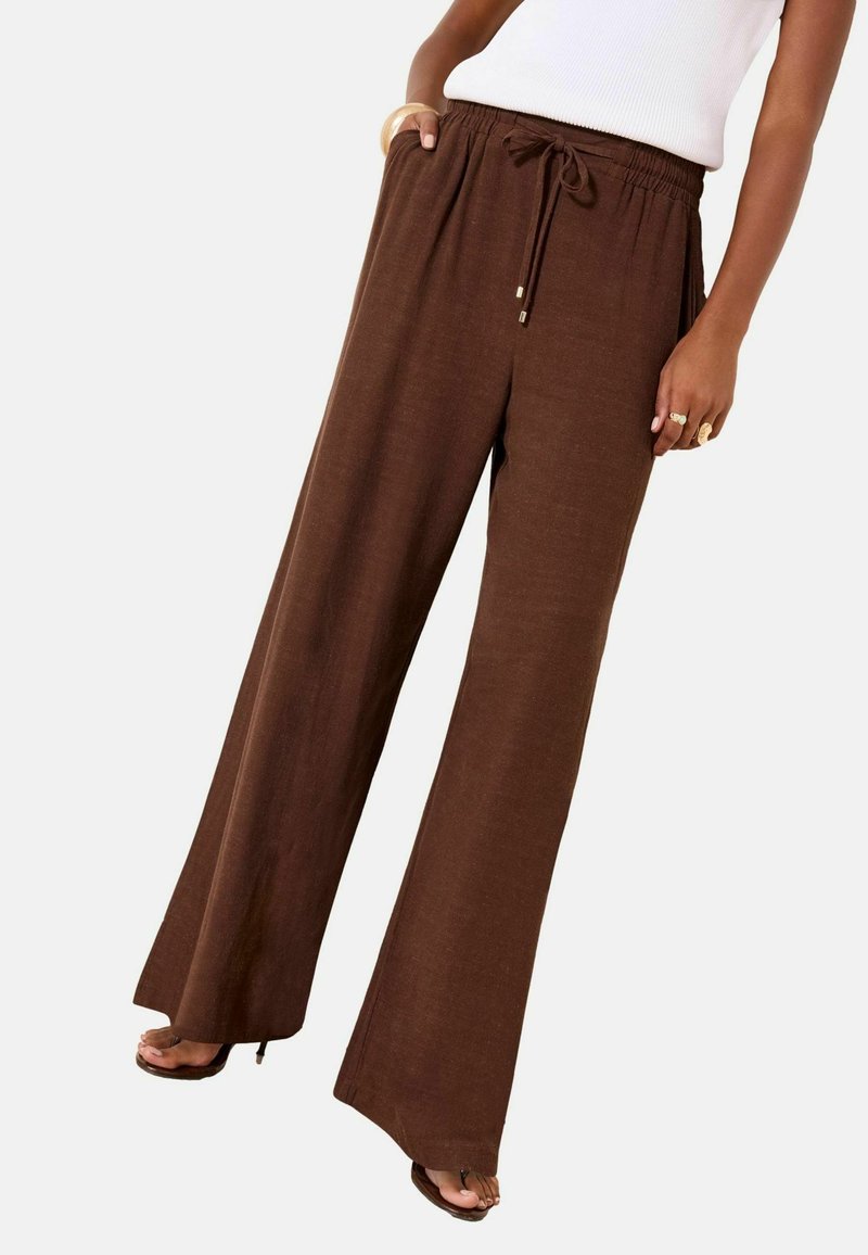 Femeie purtând pantaloni maro cu talie cu șnur și croială largă, top alb fără mâneci, inele aurii și pantofi cu toc și degetele goale, pozând cu o mână în buzunar.