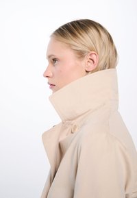 Beige trench coat with a high collar, featuring a button detail on the side. Smooth fabric, structured design, and minimalistic style.