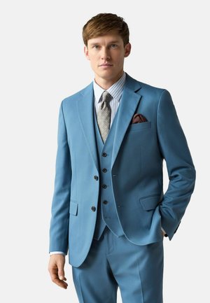Fiatal férfi világoskék háromrészes öltönyben, csíkos ingben, mintás nyakkendővel és barna zsebkendővel egyszínű háttér előtt.