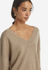 Beżowy sweter z dekoltem w serek, wykonany z miękkiego materiału, z luźnym krojem i prążkowanymi akcentami przy dekolcie i rękawach.