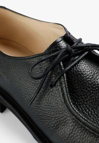 Zapato de cuero negro con una superficie texturada, punta redonda y diseño de cordones. Presenta cordones negros delgados y un interior forrado en beige suave.