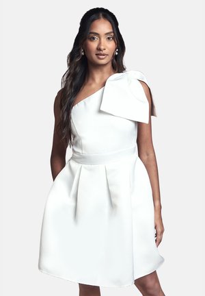 Frau mit langen dunklen Haaren, die ein weißes Ein-Schulter-Kleid mit einer großen Schleife auf der Schulter vor einem schlichten Hintergrund trägt.