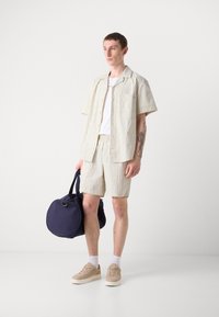 Lightweight, striped short-sleeve shirt and matching shorts in cream and beige, paired with beige sneakers and a navy duffel bag.