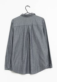 Long-sleeved shirt in grey fabric featuring a small geometric pattern, button closure, and pleats at the back. Hangs on a black hook.