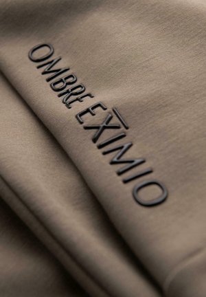 Detailní záběr na taupe látku s reliéfním nápisem "OMBRE EXIVIMIO" v čern�ých vystouplých písmenech.