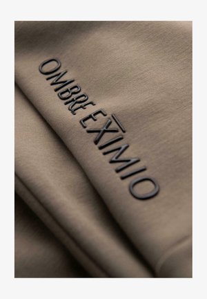 Nahaufnahme eines taupefarbenen Stoffes mit dem eingeprägten Schriftzug "OMBRE EXIVIMIO" in schwarzen, erhabenen Buchstaben.