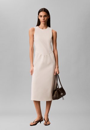 Donna che indossa un vestito midi beige senza maniche e tacchi neri con cinturini, tiene una borsa marrone scuro e si trova davanti a uno sfondo grigio uniforme.