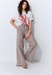 Balti grafikos marškinėliai su raudonu atspaudu, kuriame pavaizduotas figūra. Rudos ir baltos languotos plataus pūsto stiliaus kelnės su raišteliu juosmens srityje. Butai rudi sandalai.