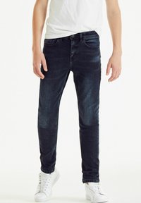 Sötétkék skinny farmernadrág klasszikus öt zsebes dizájnnal és enyhe kopással. Fehér sportcipővel párosítva.