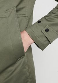 Cappotto verde oliva con superficie texturizzata, dotato di una grande tasca frontale e dettagli di polsino con bottoni.