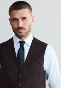 Tmavohnedý formálny vest, vyrobený z textúrovanej látky, nosený cez bielu košeľu s dlhým rukávom, doplnený čiernou viazanou kravatou so vzorom. Viditeľný jednoduchý detail vrecka.