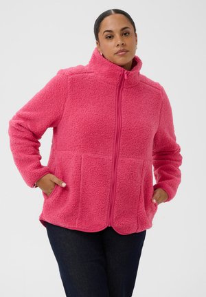 Femme aux cheveux foncés portant une veste polaire rose vif avec une fermeture éclair à l'avant et les mains dans les poches, debout devant un fond uni.