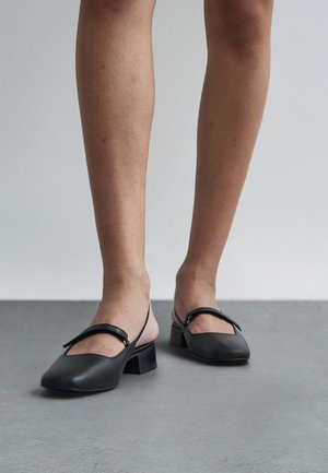 Persona valkā melnas ādas sandales ar zemu taisnstūrveida papēdi, stāvot uz pelēkas grīdas pie baltas sienas.