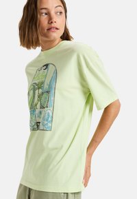 Camiseta de algodón verde claro con un diseño gráfico colorido que incluye elementos botánicos y patrones geométricos en tonos azules y negros.