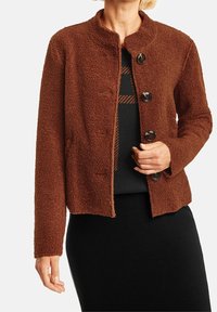 Brun texturerad jacka med rund halsringning, prydd med fyra stora knappar. Tyget ser mjukt och mysigt ut. Bärs över en svart topp.