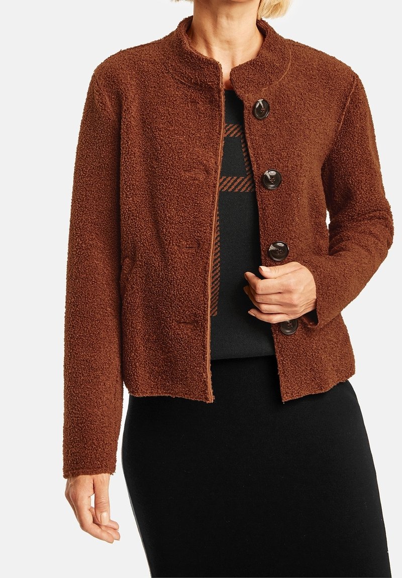 Brun texturerad jacka med rund halsringning, prydd med fyra stora knappar. Tyget ser mjukt och mysigt ut. Bärs över en svart topp.